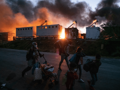 Refugees and asylum seekers walk with their possessions as a fire burns behind them in Moria Reception and Identification Centre.